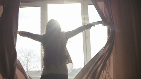 Attractive young woman opens curtains in slow motion is looking at the sunrise standing near the window in her home and enjoys of city view from height self isolation, infection, coronavirus, home