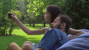 Young couple smiles people sitting on a chair in the park and makes selfie with different things.