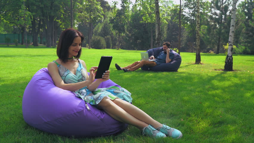 Young attractive brunette girl with bright accessories sitting in a chair and ottoman in the Park Turns the page on the tablet. Smiling, happy.
