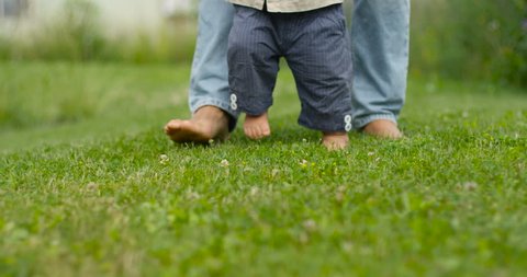 Father learns child walk outdoors. Baby first steps on grass, slow motion.
