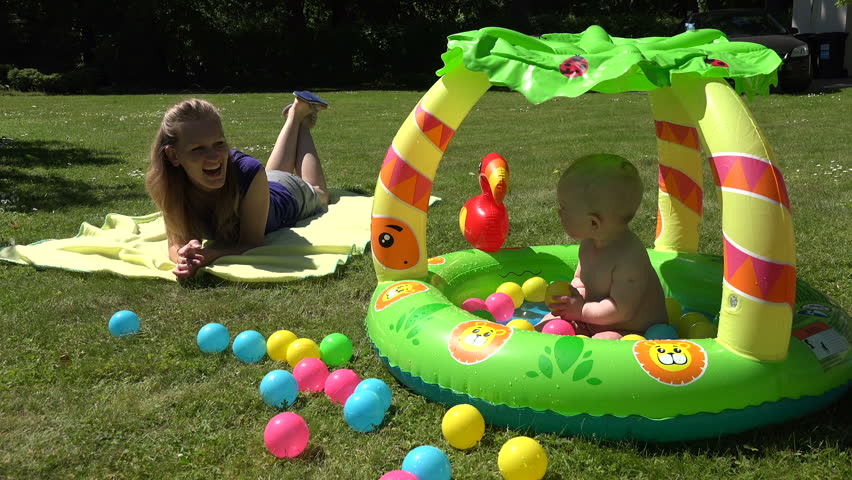 Mother play with cheerful baby splash water in inflatable pool full of colorful balls on garden meadow. Static shot.
