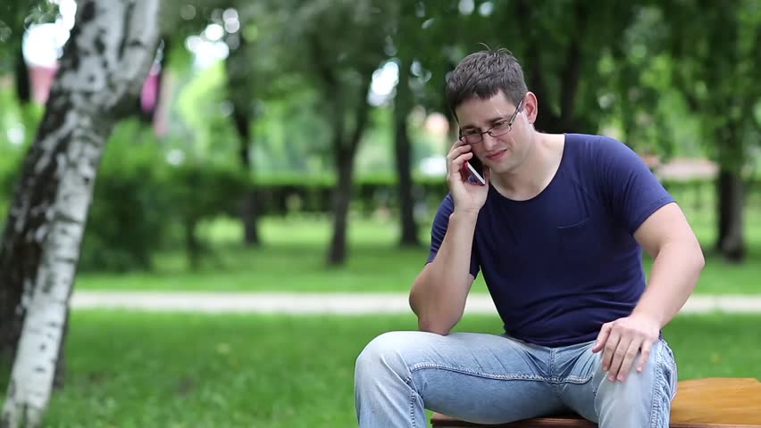 Man communicates via smartphone. Adult man sits on the bench in city park and speaks on smartphone. Man sits on the bench in public garden and uses cell phone Royalty-Free Stock Footage #18934235