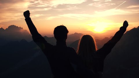 Achievement concept, edited - Embraced couple raises their arms celebrating successful climb on the mountain. Silhouette of people in beautiful sunset