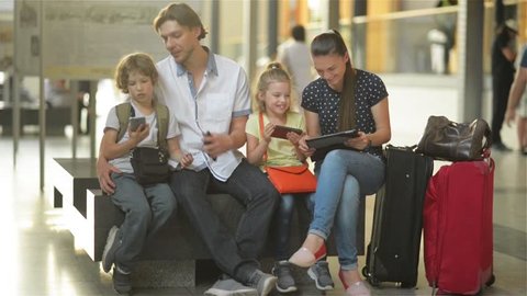 Family waiting for your flight or train and use the tablet and phones while sitting in waiting room of airport or railway station.