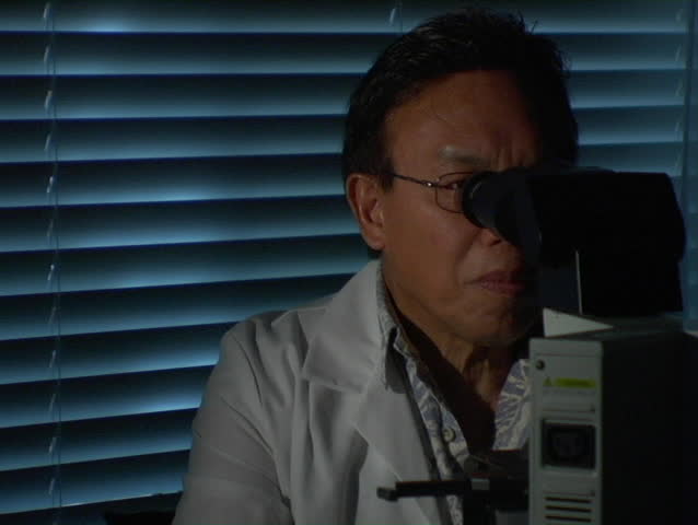 A man in a white coat looks up from a microscope.