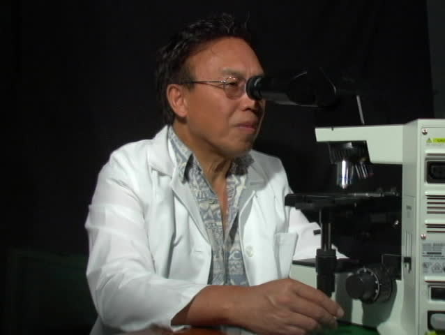 A man in a white coat looks up from a microscope.