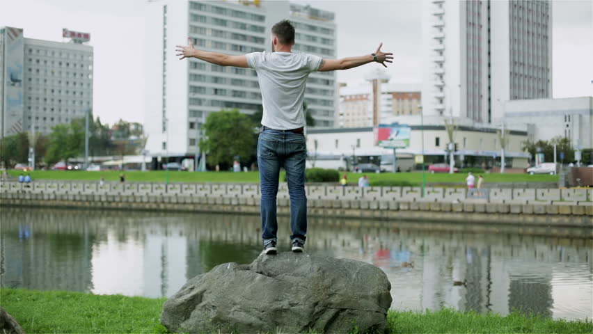 Attractive young man standing on a big stone by the river. The guy in jeans and blue t-shirt is holding up his hands. The bearded man is turning around standing on a rock. City buildings.