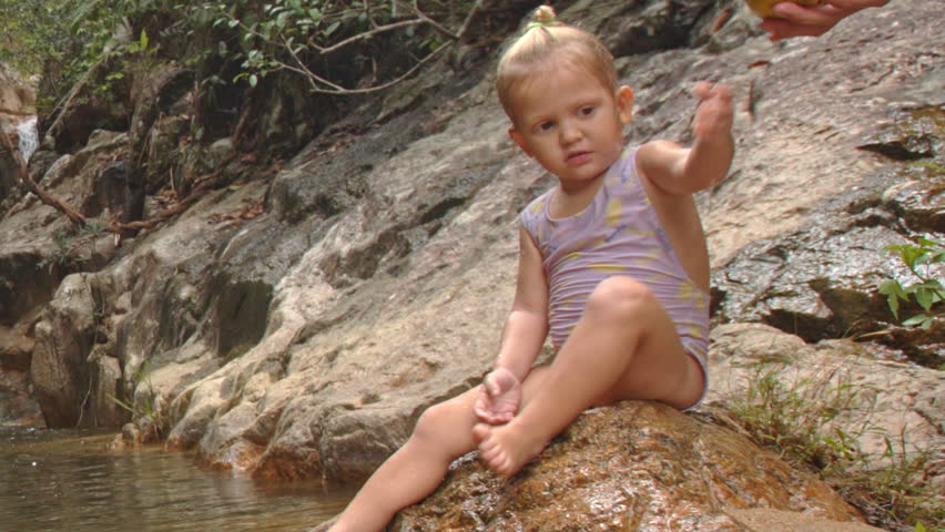 closeup little blond girl in swimsuit sits cleans foot on stony bank of mountain river with waterfall