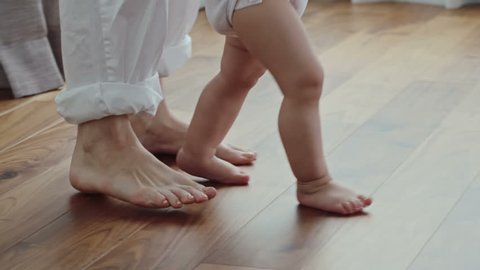 Side view of barefoot legs of woman walking on the floor behind little baby in diaper