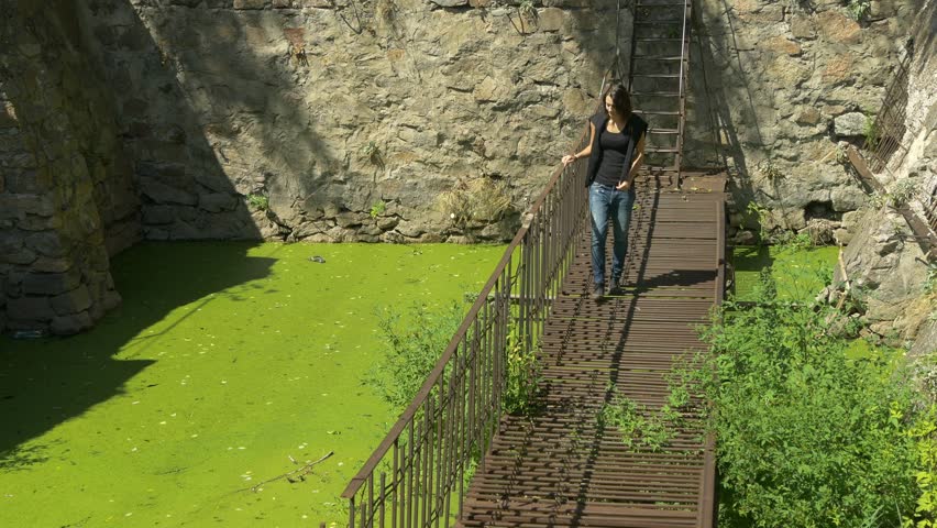 The young beautiful girl walks on a thin ruined rusty metal bridge over the dirty old green water. Hydroelectric dam across the river. High vintage stone wall in the background.