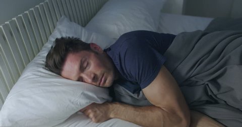 Sleeping man turns towards light while dreaming in a double bed with soft ambient light.  Camera move on jib arm.  Side view, medium shot.