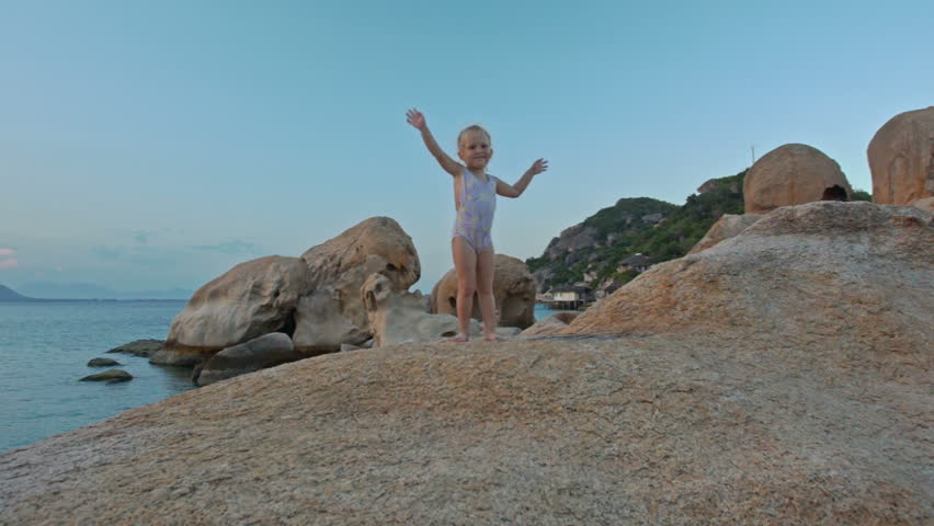 closeup little girl in swimsuit stands on top of large bare rock on beach waves hands against sea stones sky at sunset