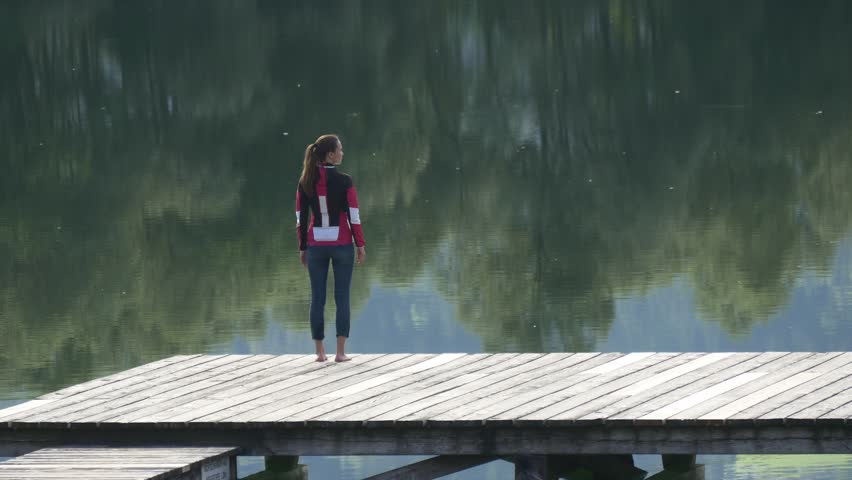 Thoughtful woman standing on dock and looking at clear reflection of trees on the water