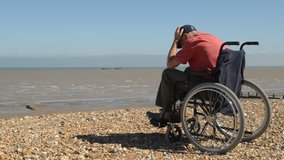 Disabled man in a wheelchair on the beach, facing the sea on the right hand side of frame.