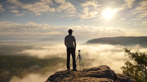 Photographer in grey t-shirt and red cap preparing tripod on cliff. Dreamy fogy landscape, blue misty sunrise in a beautiful valley below