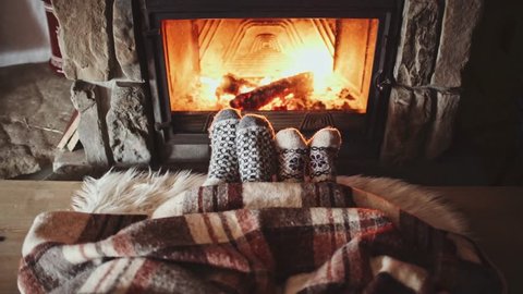 Couple Feet in Wool Socks by the Cozy Fireplace, 4K. Man and Woman sitting under the blanket, relax by warm fire and warming up their feet. Close up. Winter and Christmas holidays concept. 