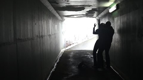 Silhouette of a criminal adult male attacks a young adult woman from behind in an underground dark alleyway tunnel. Violence against women concept with copy space. 