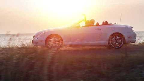 Young Fun Couple Driving Convertible At Sunset At Sea Front Beach Woman Waiving Hands Dancing Joy Adventure Flyover Drone Shot