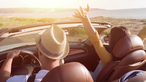 Cute Young Couple Driving At Sea Front Woman Enjoying Waiving Arms In The Air In Convertible Car Wind Blowing Hair Summer Fun Vacation Adventure Concept