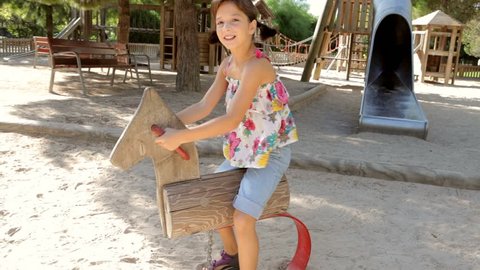 Girl child sitting on a wooden swinging horse in a park playground, swinging and smiling on a sunny day on holiday, outdoors. Activities fun games lifestyle, kids recreation vacation time, exterior.