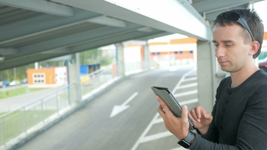 Outdoor portrait of modern young man with digital tablet in the street. The man in glasses with backpack sitting on the bridge. Bottom drive cars