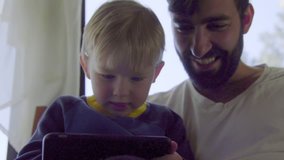 From a low close angle, a young boy and his father smile and laugh as they watch a movie on a cell phone