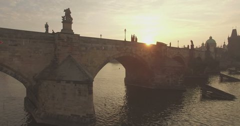 Aerial camera flies fast along the Charles Bridge in Prague as the sun rises behind it.