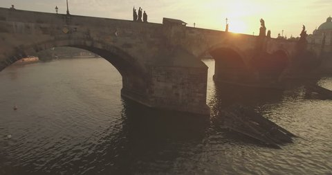Aerial camera flies backwards fast from the Charles Bridge in Prague as the sun rises behind it