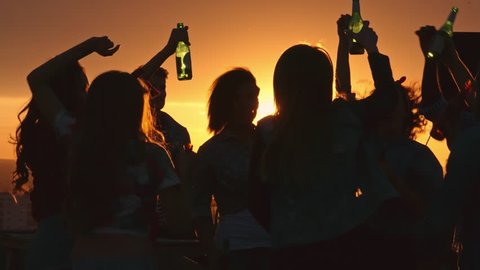 Silhouettes of young people toasting with beer bottles and dancing with raised arms to the music played by dj at rooftop party during beautiful city sunset