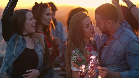 Beautiful young couple with sparklers slow dancing and kissing passionately during rooftop party at sunset, multi-ethnic group of young people dancing around them