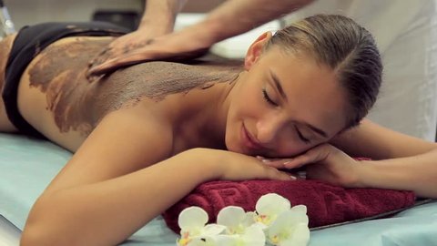 Masseur applied uniformly brown mass on a woman's back. He uses a scrub while relaxing in the spa. Nearby lie fresh flowers. 