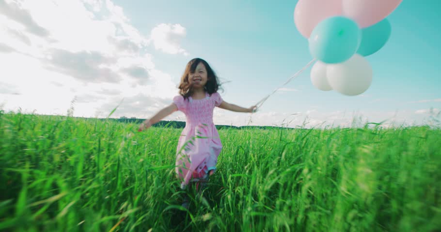 Little Asian girl in a dress running through green wheat field with balloons in hand, slow motion