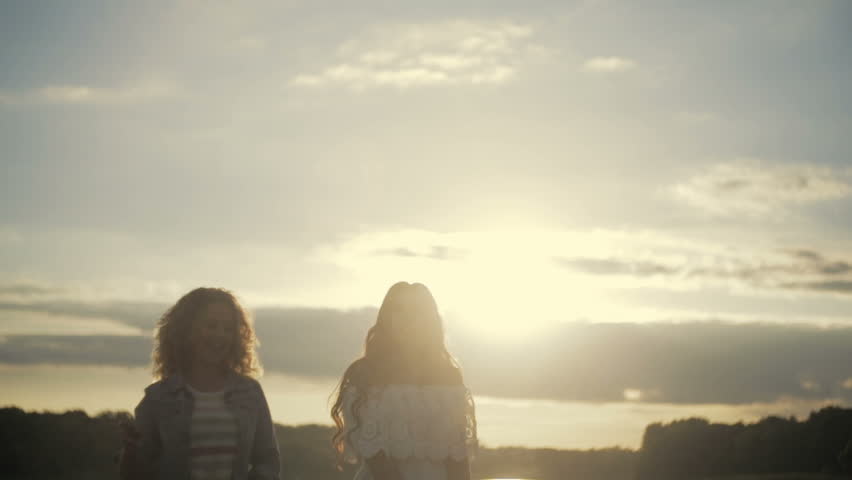 Young girls are having a little party by the lake. Dancing, singing in bright sunshine. Casual wear, elegant white dress. Summer sunset. Females portrait, no people around.