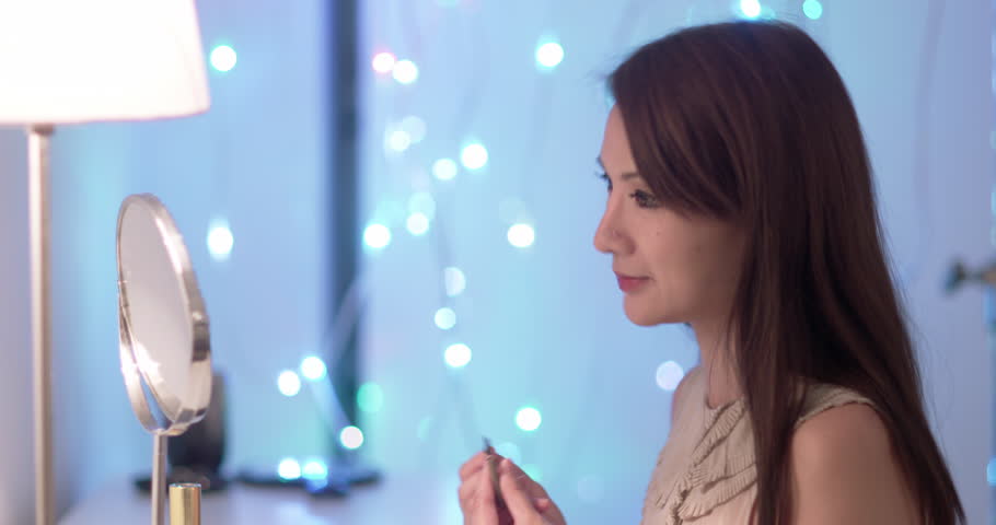 Attractive young Asian woman makes up her eye brows with a dark pencil while sitting at dressing table.  Close up side view with soft focus highlights in background.