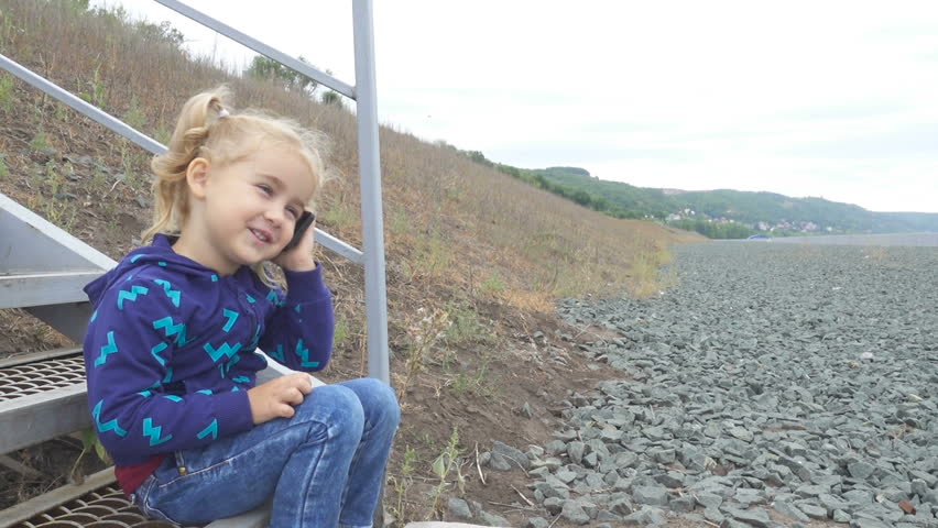 Pretty little girl talking on phone sits on the stairs on the banks of the river. 