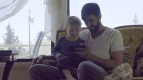 Medium shot in nice living room of young bearded father showing adorable son sitting on his lap something on a smart phone
