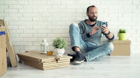 Young man chatting on smartphone sitting on floor at his new home 
