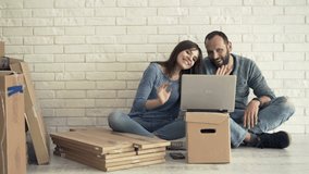 Young couple chatting on laptop sitting on floor at their new home 
