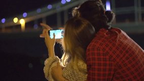 Beauty happy young couple dating, taking picture of beautiful night city on smart phone, hugging and kissing together. Girlfriend and her boyfriend walking on night streets, enjoying night view. Love