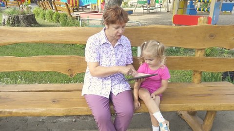 Grandmother Teaching Granddaughter  How to Use a Tablet PC. Sitting on a bench in the park sunny summer day. 