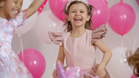 Exhilarated little girls dressed like princesses jumping on couch and holding gift boxes at birthday party in slow motion