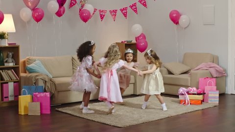 Four beautiful little girls dressed like princesses dancing in circle in decorated kids room at birthday party