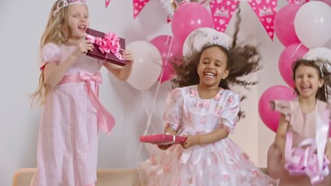 Four little girls dressed like princesses jumping on sofa and holding gift boxes at birthday party in slow motion