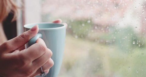 Young Woman Enjoying her morning coffee or tea, Looking Out the Rainy Window. 4K DCi SLOW MOTION 120 FPS. Beautiful romantic unrecognizable girl drinking hot beverage at cozy home. Rainy Day Mood.
