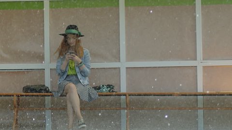 Girl sitting at the bus stop in the rain and texting with mobile phone . People running in the rain with umbrellas. Shot on RED EPIC Cinema Camera in slow motion.