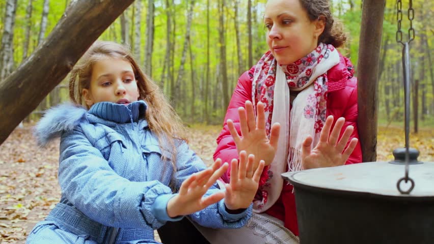 Woman and girl warm hands near kettle and smolder log