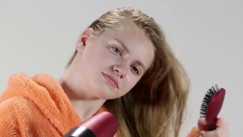 Face of blonde teenage girl wearing orange bathrobe blow drying her wet long hair with hairdryer after shower in bathroom closeup