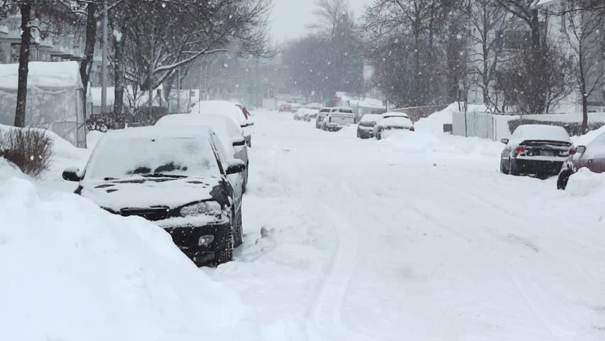 Residential street during a snowstorm. The cars are  covered in snow.