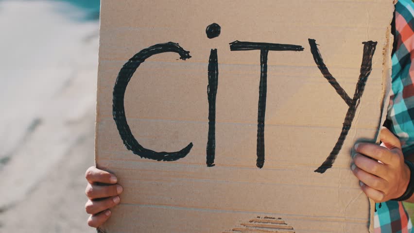 Boy hold cardboard plate with sign city hitchhiking at road. Cars. Traveler. Summer sunny day.
