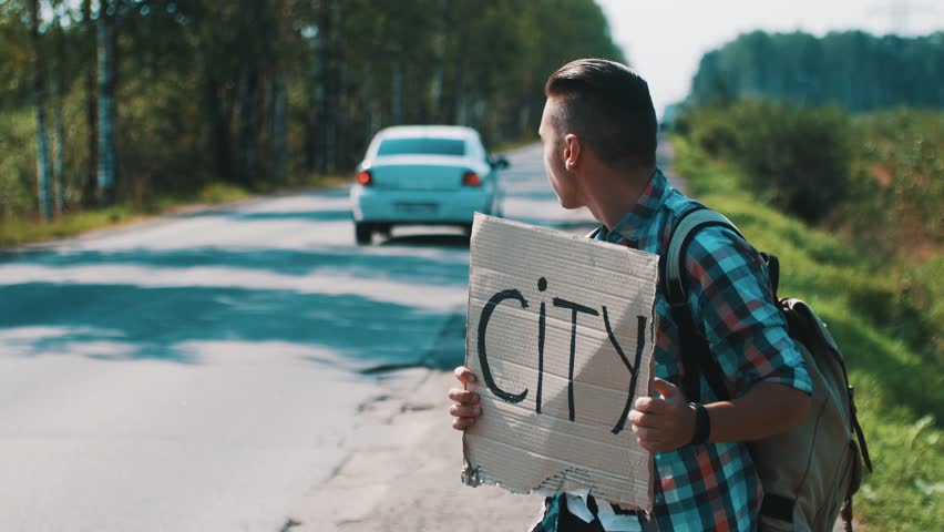 Boy hold cardboard plate with sign city hitchhiking at road in summer sunny day. Cars.